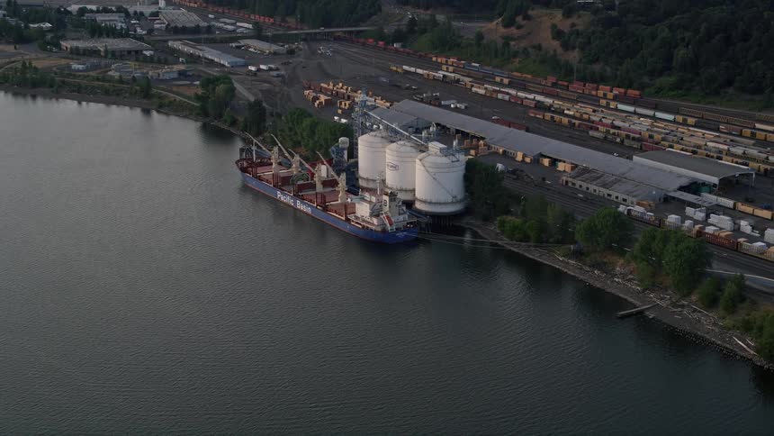 Aerial footage of an oil tanker approaching a silo and train yard beside the Willamette River in North Portland Oregon during sunset