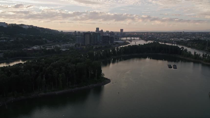 an aerial view of Ross Island in the Willamette River with the stunning metropolitan skyline of Downtown Portland Oregon in the distance as the sun sets over the city during a summer evening