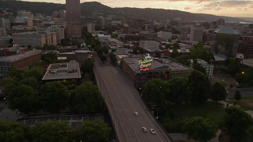 Aerial view of downtown Portland Oregon at sunset approaching the iconic White Stag Sign as the Willamette River flows below showcasing the city's historical landmarks and high-rise buildings