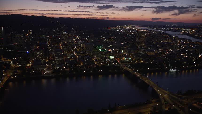  aerial footage showcasing the stunning high rise buildings bridges and cityscape of downtown Portland Oregon during twilight with partly cloudy skies and the beautiful Willamette River flowing through the city