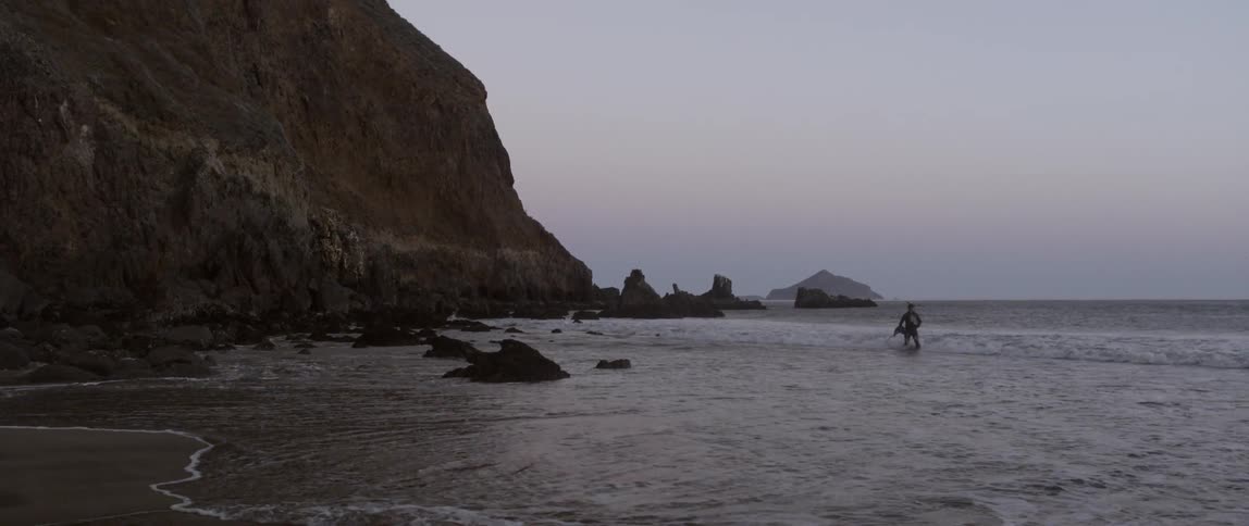 A man is walking in the water of a beach as waves splash on him