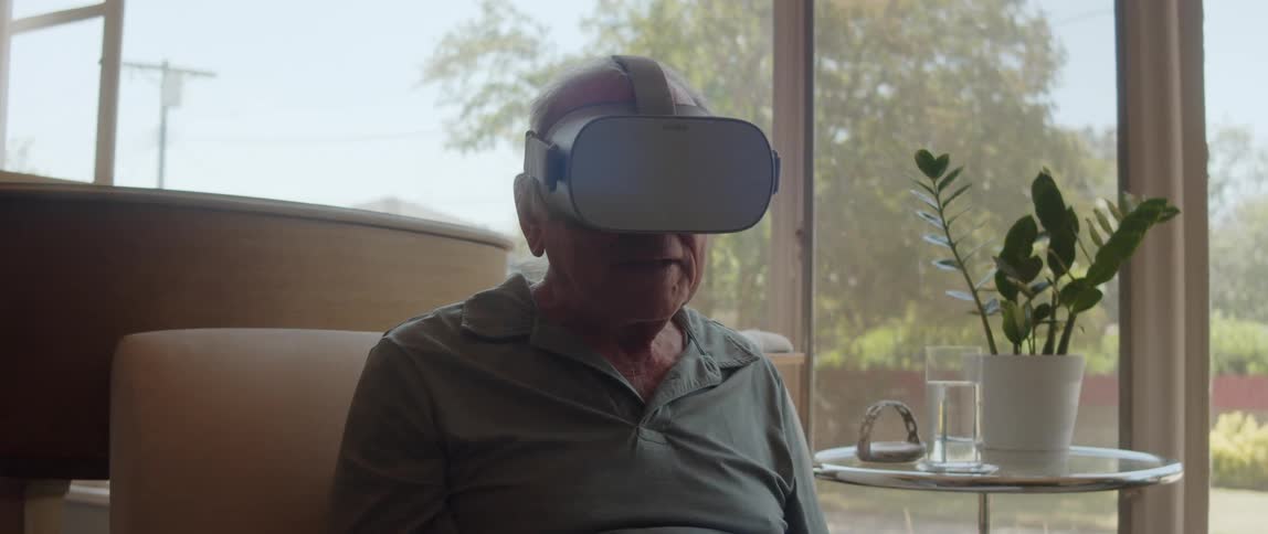 An elderly man wearing a vr headset while sitting in the living room next to a table with a flower plant