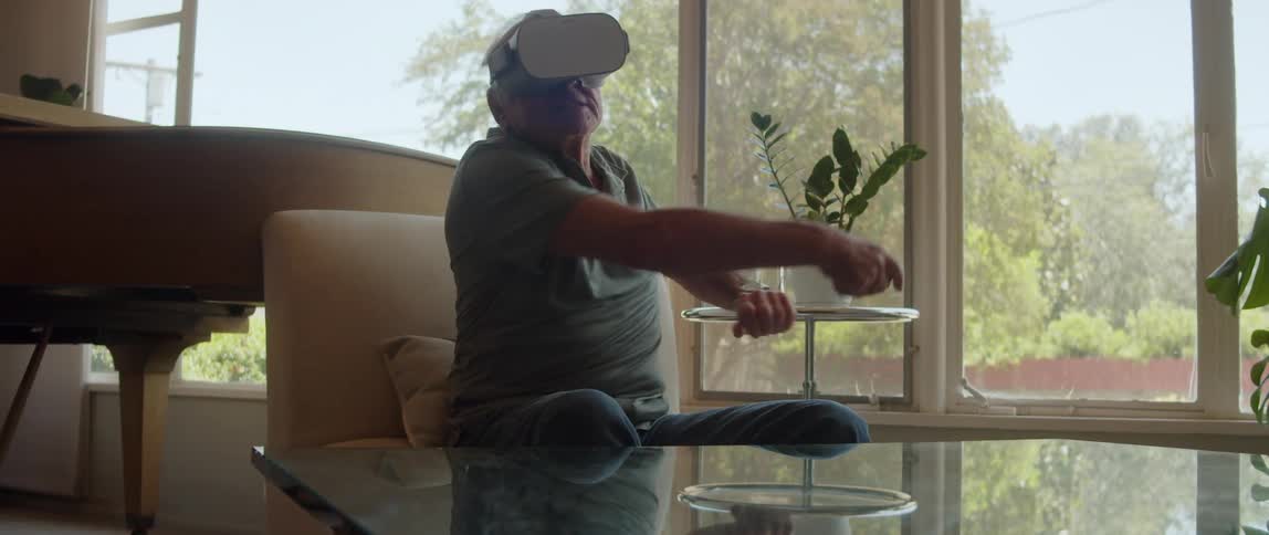 An ederly man playing a game while wearing a vr headset seating next to a glass table