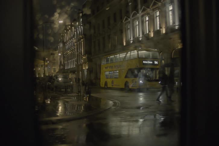 From the car windows perspective a doubledecker bus is stationary on the roadside beside a building pedestrians clutch umbrellas tightly as they navigate the raindrenched street