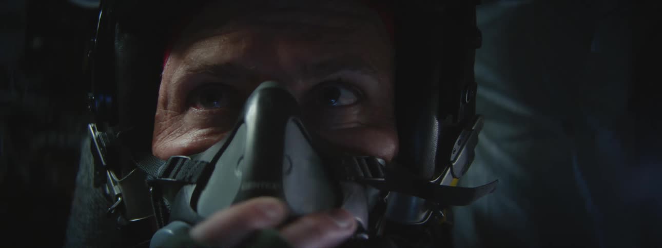 A male airplane pilot is wearing a helmet and adjusts his oxygen mask while in the cockpit of the plane