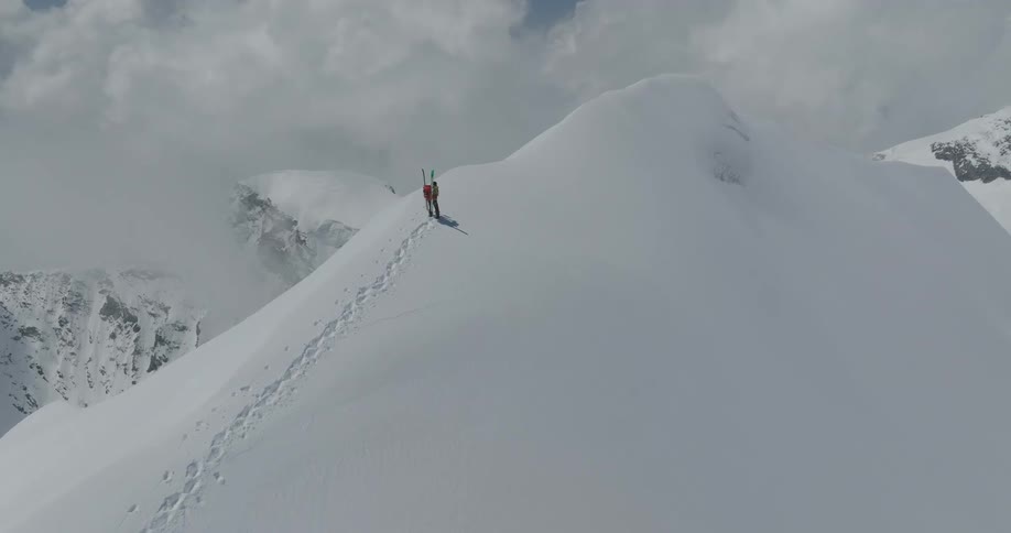 Two individuals stand atop a snowcovered mountain leaving behind their footprints along the path etched into the snowy terrain