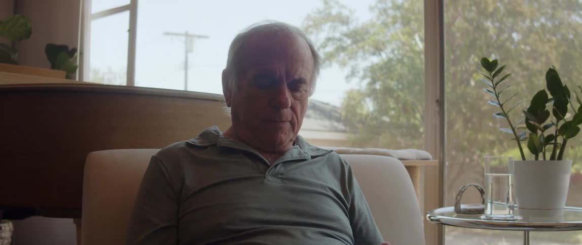 An elderly man is sitting indoors and texting on his cellphone
