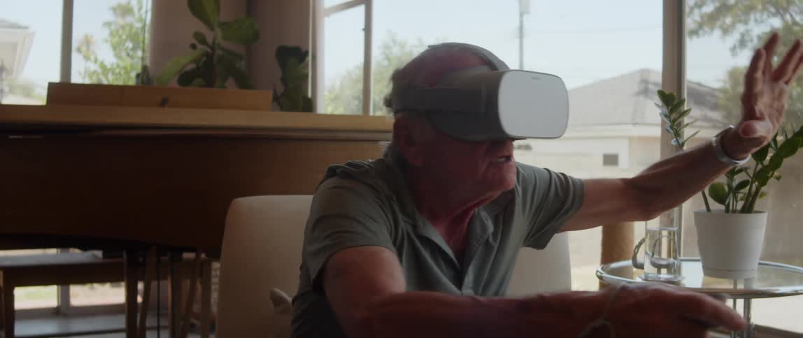In the living room an elderly man sits immersed in a virtual reality world wearing a headset and engrossed in playing a video game