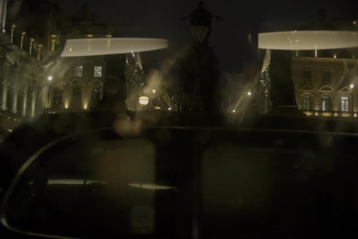 In the dimly lit cityscape of the night cars glide along the streets while a solitary figure stands on the sidewalk