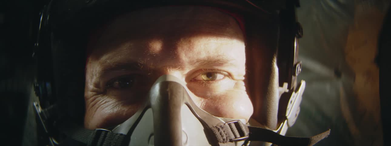 An aircraft pilot wearing a helmet and oxygen mask sits in the cockpit a shadows is cast upon his face