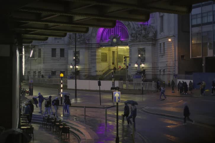 Pedestrians are strolling along the wet streets at night with some individuals carrying umbrellas to shield themselves from the rain