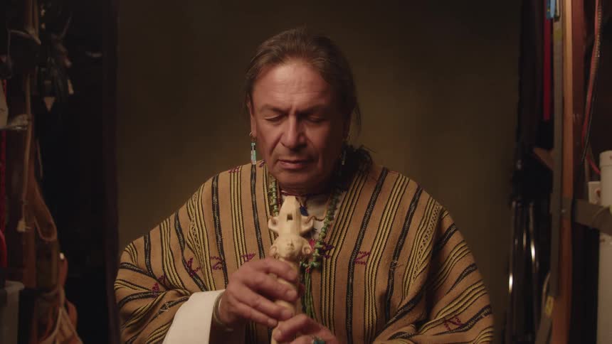 A native american man sits down prays and prepares to play a decorated flute