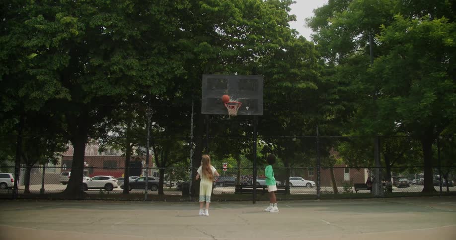 Two kids play basketball in park across street from building with parking lot