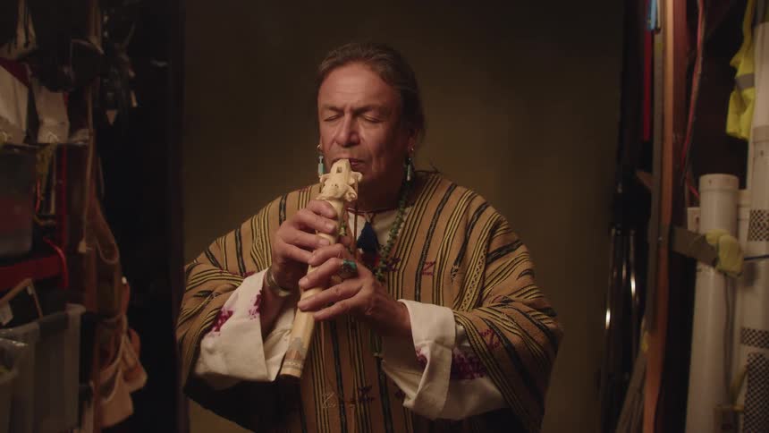 Portrait of an indigenous man sitting and blowing a flute while a photography flash goes off