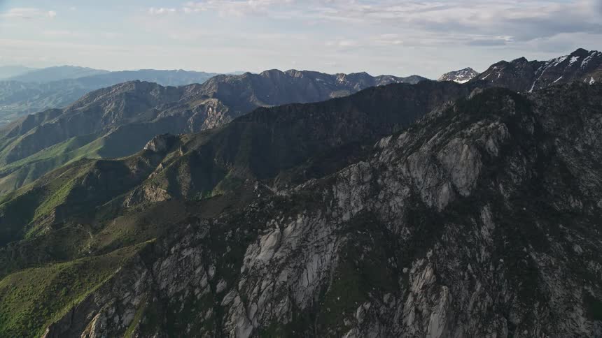 Approaching the snow-capped peaks of the Wasatch Range in Utah's Southwest United States during a partly cloudy summer day capturing the breathtaking mountain ridges and natural wilderness from an aerial perspective