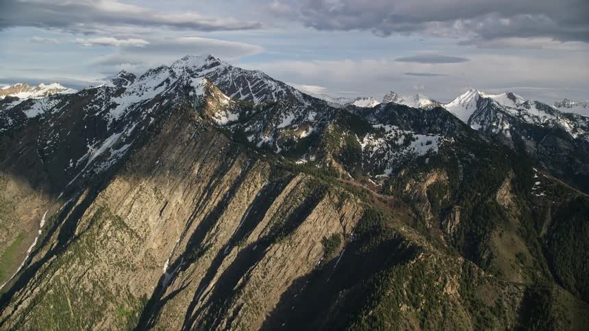 Approaching the snow-capped peaks of Wasatch Range in Utah during summer pan left to reveal the majestic mountain landscape with partly cloudy skies and white snow mountains
