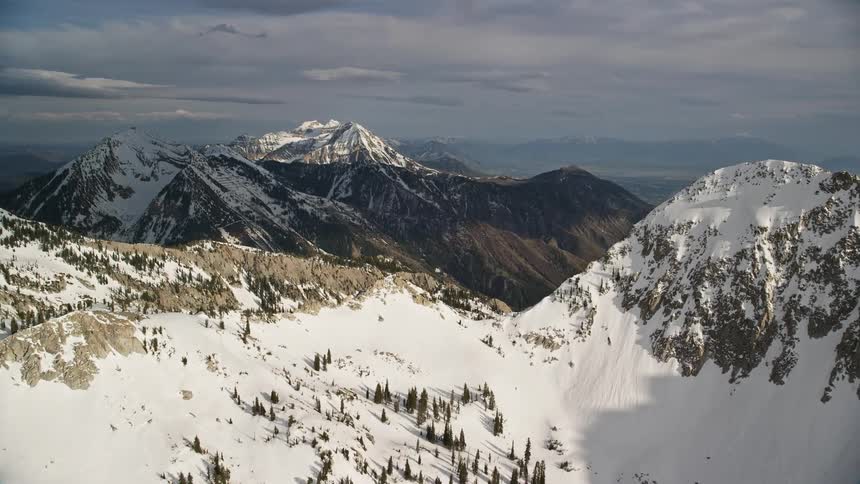 Approach a snowy mountain peak in Utah's Wasatch Range surrounded by coniferous woods during a partly cloudy winter day