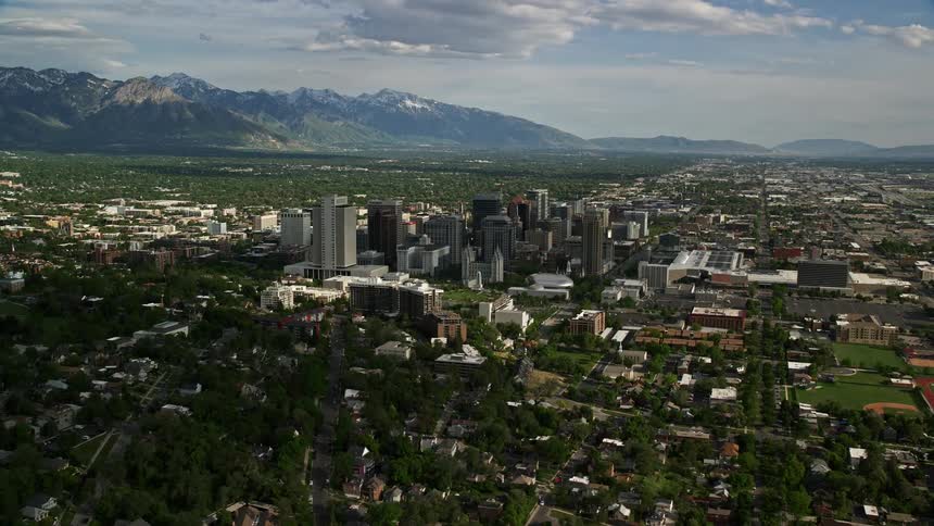 Aerial flyby of Downtown Salt Lake City Utah showcasing city buildings homes sports fields and the stunning mountainous backdrop
