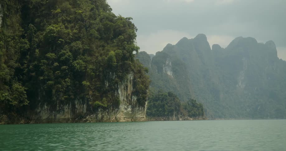 A river flows with rocky banks where trees take root and grow atop the stones in the distance a mountain rises against a sky shrouded in dark clouds