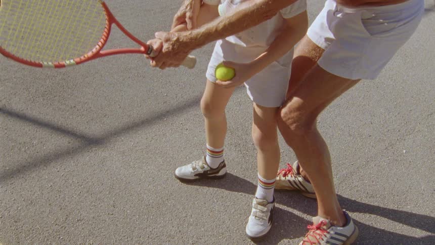 A man is teaching his young son how to stand and hold the tennis racket and hit the ball