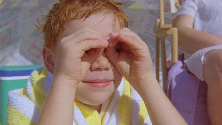 Child with both hands over their eyes in a binoculars gesture and carrying a beach towel on their back