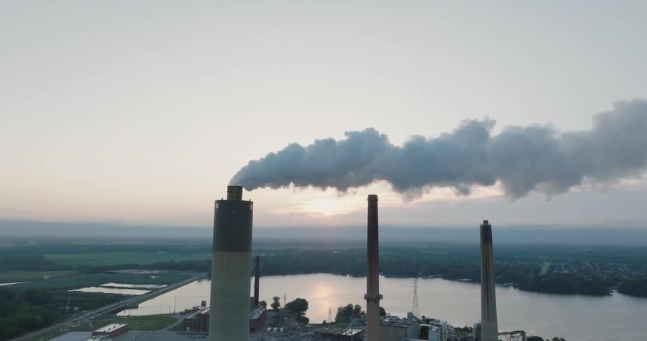 Smoke and steam rise from a tall chimney into clear skies with the sun on the horizon