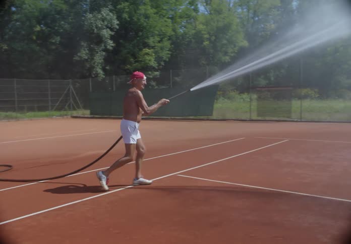 Elderly man watering a tennis court with a hose