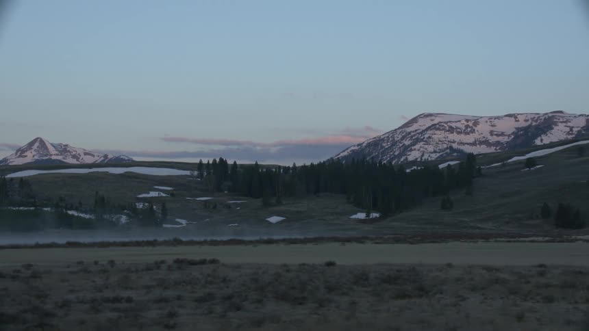 A field with trees and patches of snow and mountains filling the horizon