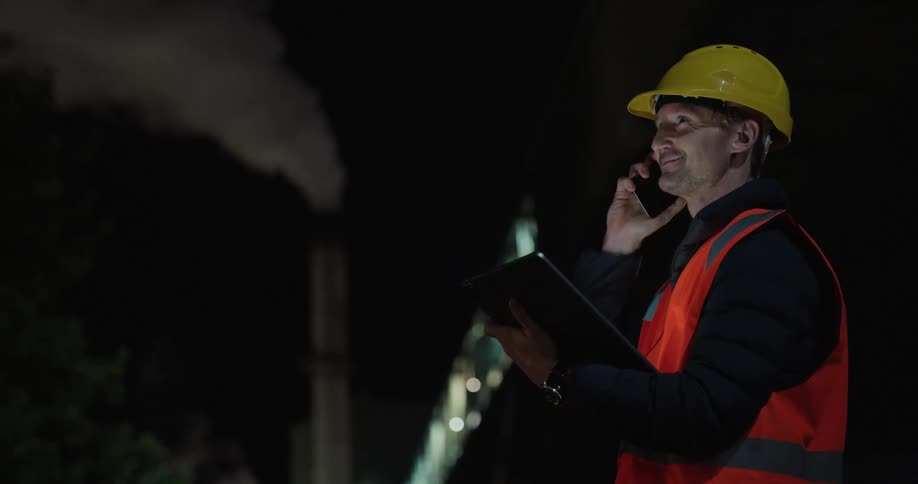 A man wearing a hard hat and vest while talking on the telephone at night looking at a tablet in his hand