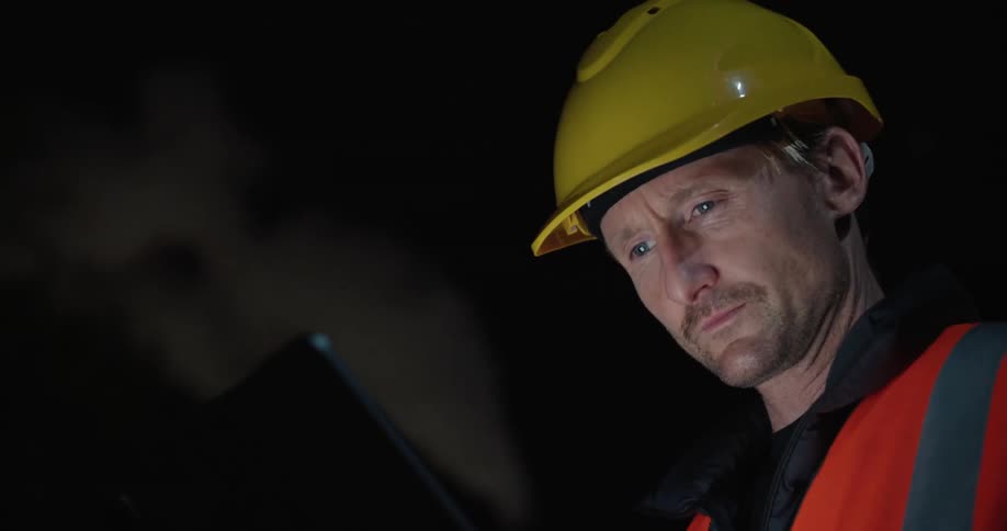 Worker wearing hardhat consults tablet at night