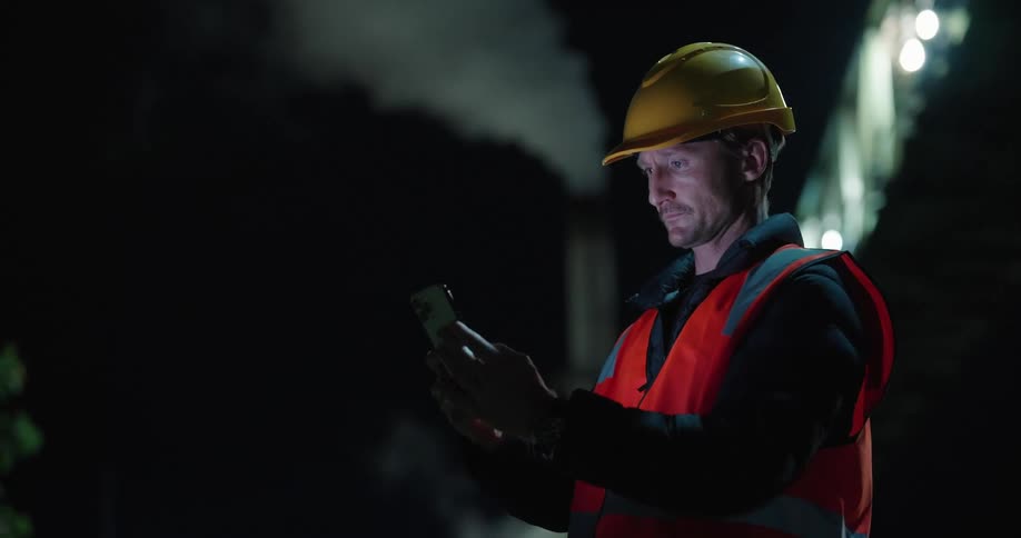 A man in a builders suit and helmet fiddling with a smartphone with a glowing screen