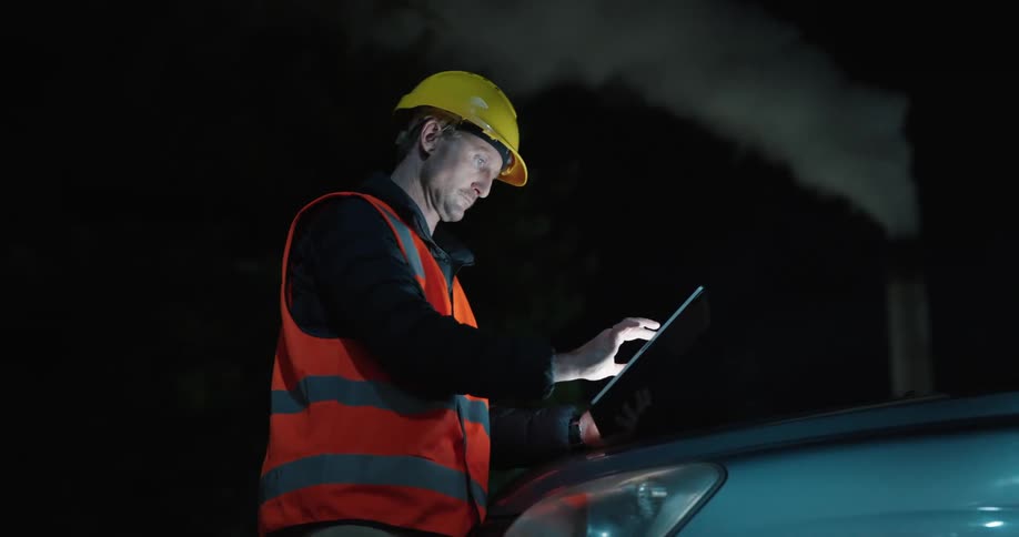 Man wearing a protective helmet and highvisibility clothing using a tablet and leaning on the hood of a car