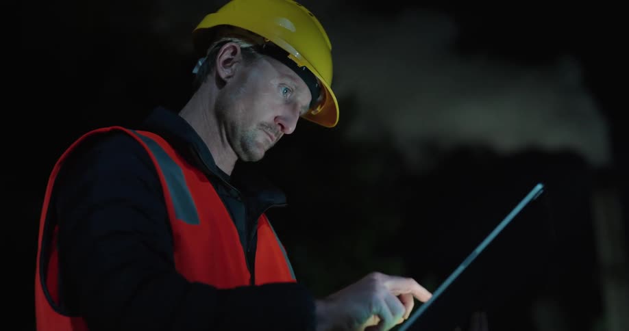 A man in a yellow helmet and orange vest using a tablet with the screen glowing and then he looks to the side
