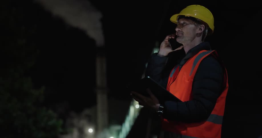 A industrial plant worker is talking outside on a smart phone at night