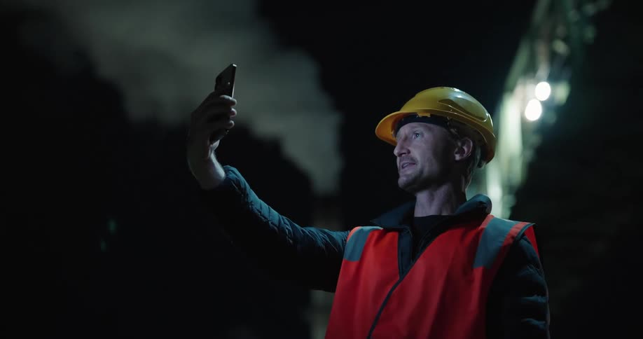 A man in a helmet and builders clothing is smiling with a cell phone on his face