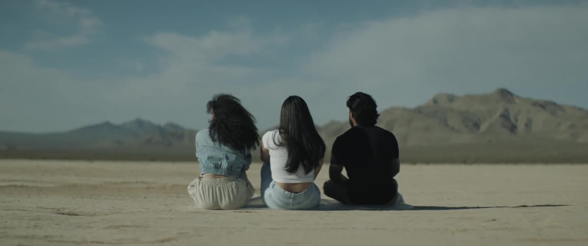 Three people sit on the ground facing mountains on horizon
