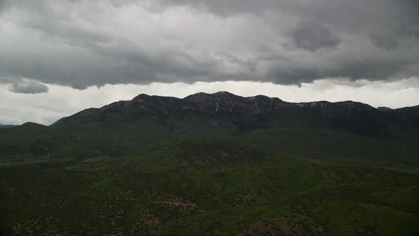 Aerial footage of Salt Creek Peak in Utah's Wasatch Range with green mountain and dark clouds on the horizon during daytime