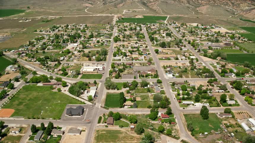 Aerial view of a small rural town nestled near farmland surrounded by rustic houses and scattered residential areas in Aurora Utah during a partly cloudy day in summer