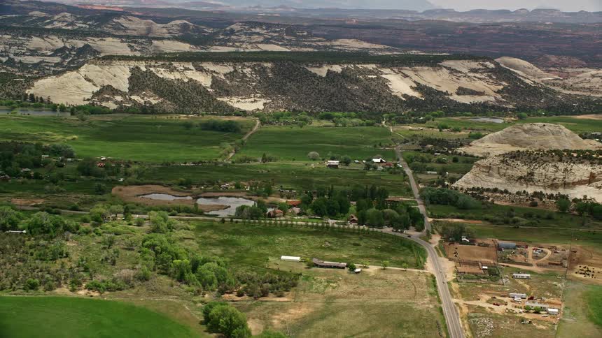 Aerial footage of a rural village nestled in a valley between mountain ridges in Boulder Utah featuring green fields a pond and partly cloudy skies