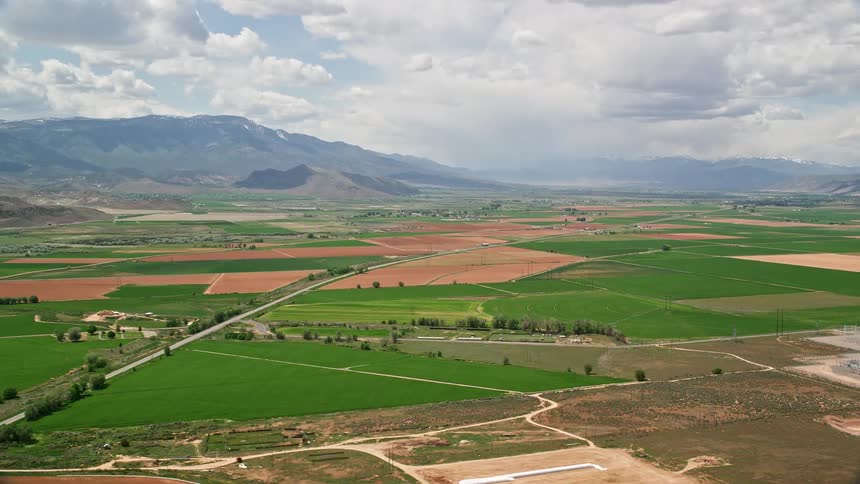 Aerial footage of a rural town in Venice Utah approaching a bend on Route 118 surrounded by mountains and farmland with partly cloudy skies