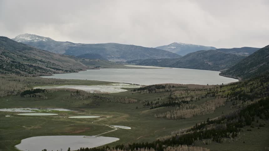  aerial footage of a vast lake surrounded by mountains and aspen trees in Fish Lake Utah with cloudy skies and a side view perspective