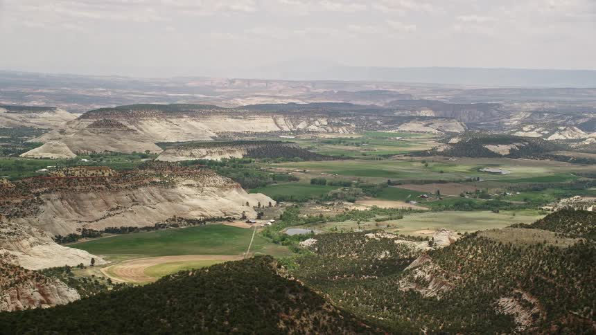Aerial footage of green fields hills covered in vegetation flying by Route 12 and tilting up to reveal the small town of Boulder Utah during the summer