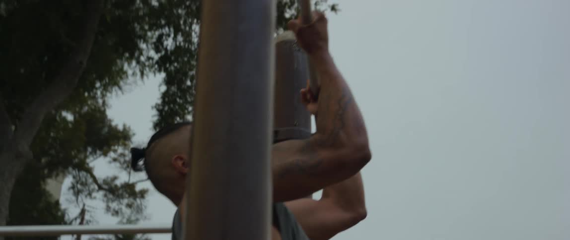 A man in an outdoor setting attempts the second repetition of a pullup but fails and angrily slaps the ground in frustration