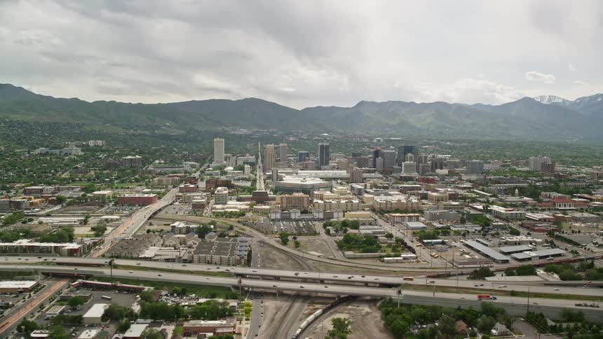 Aerial footage showcasing the bustling cityscape of Downtown Salt Lake City Utah with a backdrop of majestic mountains and light traffic on Interstate 15
