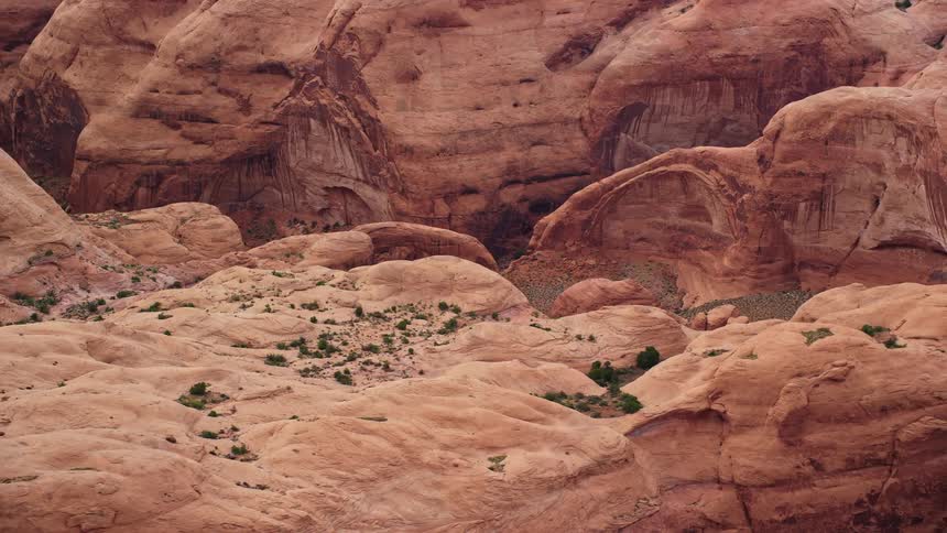  aerial footage showcasing stunning rock formations in an arid desert landscape with partly cloudy skies revealing the magnificent Rainbow Bridge National Monument in Utah as the camera flies by a mesa providing a captivating side view perspective over th