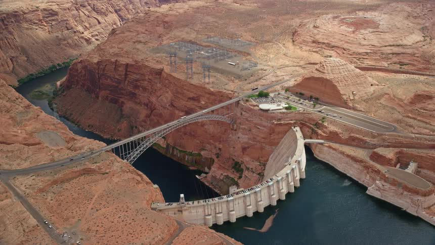 Aerial footage showcasing the Glen Canyon Dam and Bridge in Arizona with the Colorado River flowing through on a partly cloudy day during the summer in Utah