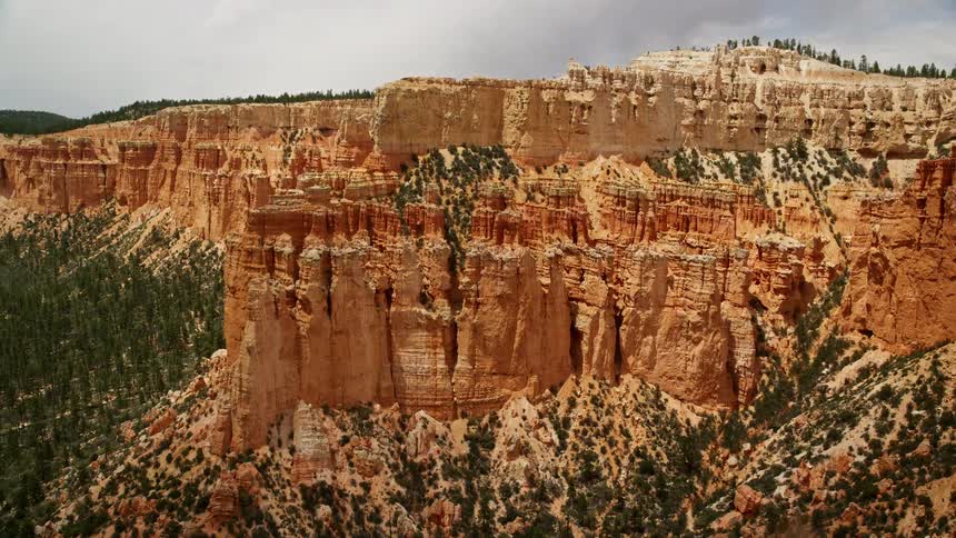 Aerial footage showcasing the stunning beauty of Bryce Canyon National Park in Utah featuring hoodoos mesa cliffs and tree-covered slopes with a focus on a tree-lined cliff and the arid rocky landscape beneath cloudy skies