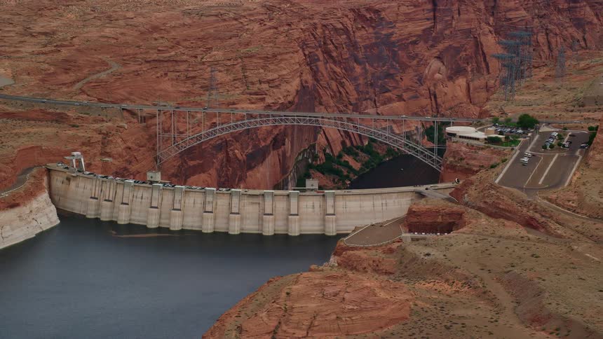 Aerial footage of Glen Canyon Dam and Bridge in red rock canyon Arizona showing the power generation and transportation infrastructure with partly cloudy skies and the Colorado River flowing beneath