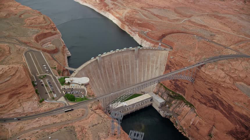 Aerial footage showcasing the Glen Canyon Dam and Bridge in Arizona circling above the Colorado River with picturesque views of the surrounding nature and arid desert landscape during a partly cloudy day in the Southwest United States
