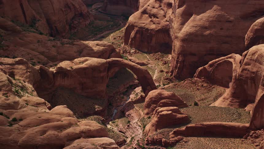  aerial footage of a stone arch in Rainbow Bridge National Monument Utah as the drone passes by in the middle of the day capturing the breathtaking rock formations and dry riverbed below